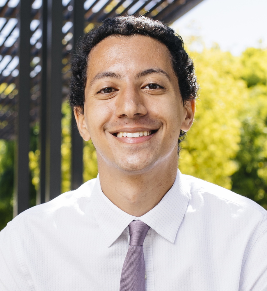 A person in a white shirt and lavender tie smiling outdoors with greenery and a pergola in the background.